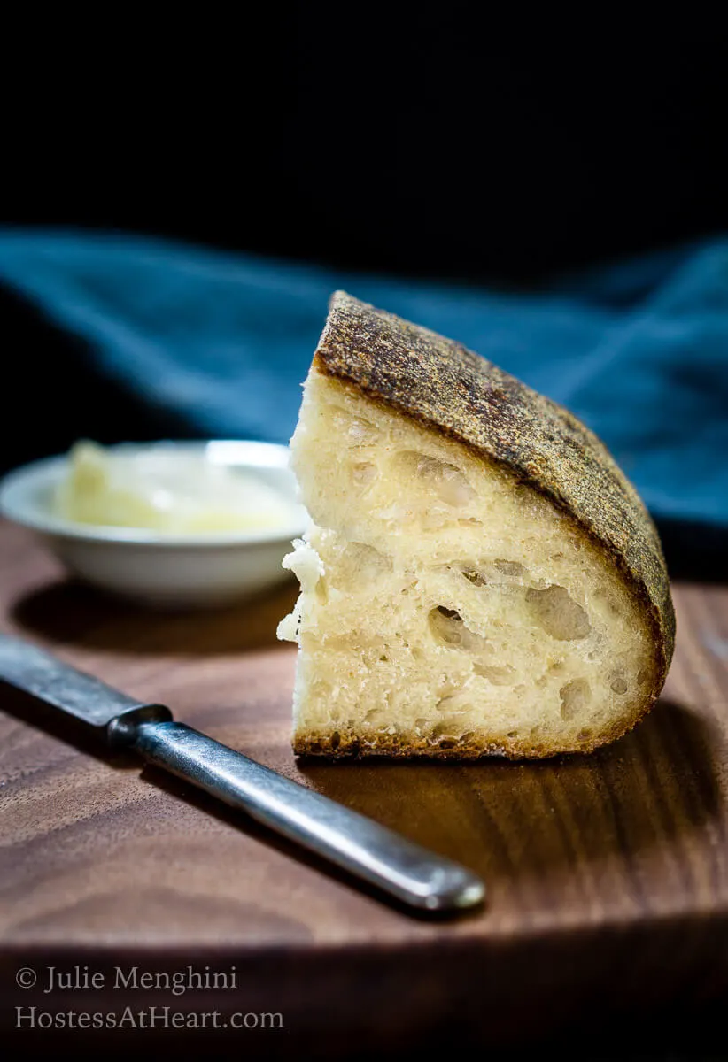 A small slice of sourdough bread sitting uprights showing the holes in the crumb over a wooden cutting bowl. A small knife and a white dish of butter sit next to the bread. A blue napkin sits in the background.