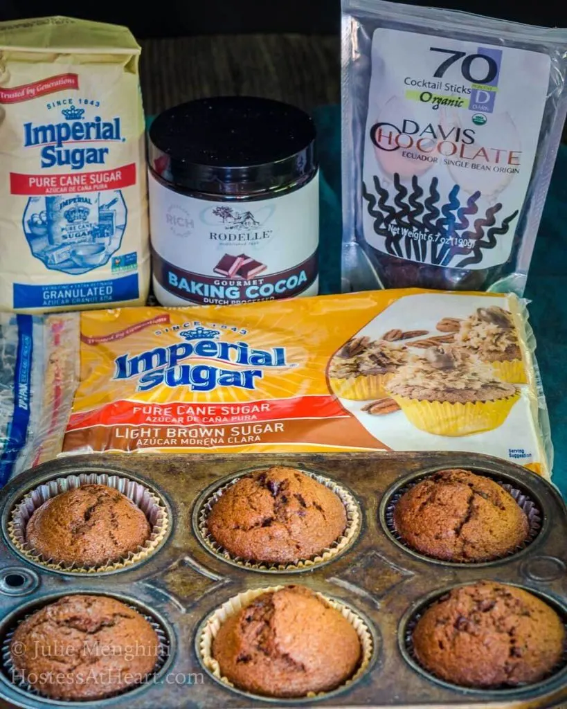 Ingredients used to make Banana Chocolate Chip Espresso muffins including Sugar, brown sugar, cocoa, and chocolate swizzle sticks. A tin of 6 muffins sit in the foreground.