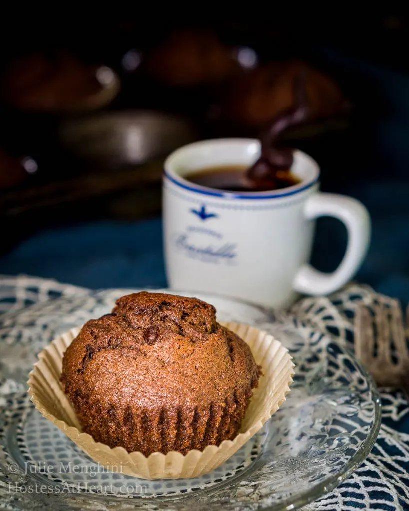 A Banana Chocolate Chip Espresso Muffin sitting in a parchment muffin paper over a lace doily.  A cup of espresso sits in the background. 
