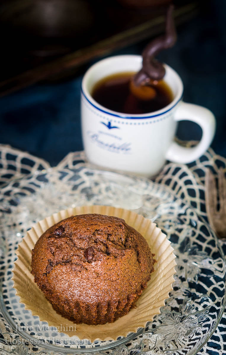 A Banana Chocolate Chip Espresso Muffin sitting in a parchment muffin paper over a lace doily.  A cup of espresso sits in the background.