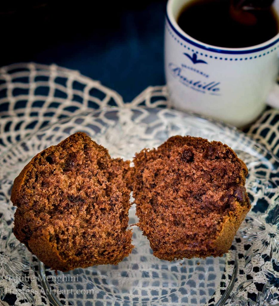 A Banana Chocolate Chip Espresso Muffin cut in half on a glass plate sitting on a lacey doily in front of a cup of espresso.