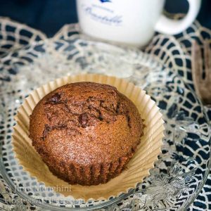 Chocolate banana muffin sitting in a parchment paper muffin paper over a clear plate. An espresso cup sits behind it.