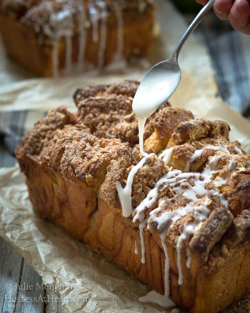 A loaf of Apple Pecan Streusel Pull-Apart Bread sitting on a piece of parchment paper. A spoon hovers over the top of the bread drizzling a white sugar glaze over the top and down the sides.