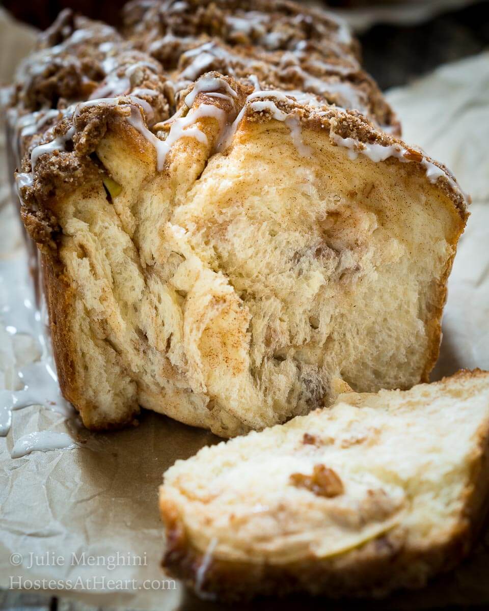 A loaf of Apple Pecan Streusel Pull-Apart Bread sitting on a piece of parchment paper with the front pieces pulled off showing a soft cinnamon and apple interior. The top is drizzled with a white glaze running down the sides.