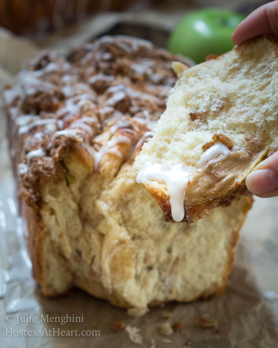 A hand holding a piece of Apple Pecan Pull-Apart Bread with a white glaze running over the front. A Loaf of the bread sits in the background.