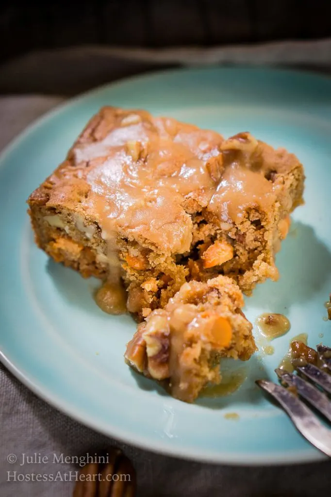 Top angled photo of a piece of butterscotch pecan bar with a brown sugar drizzle on a light blue plate. A forkful of the bar sits to the side showing the butterscotch pieces in the bar. 