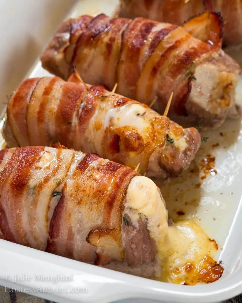 A baking dish holding three pork chop tenderloins wrapped in bacon that have cheese bubbling out of the ends.