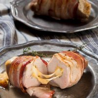 A metal plate holding a pork chop that's wrapped in bacon and stuffed with apple and cheese with the cheese pulled between the two halves sitting on a blue striped town. A second plate sits in the background holding another pork chop.