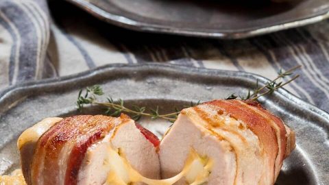 A metal plate holding a pork chop that's wrapped in bacon and stuffed with apple and cheese with the cheese pulled between the two halves sitting on a blue striped town. A second plate sits in the background holding another pork chop.