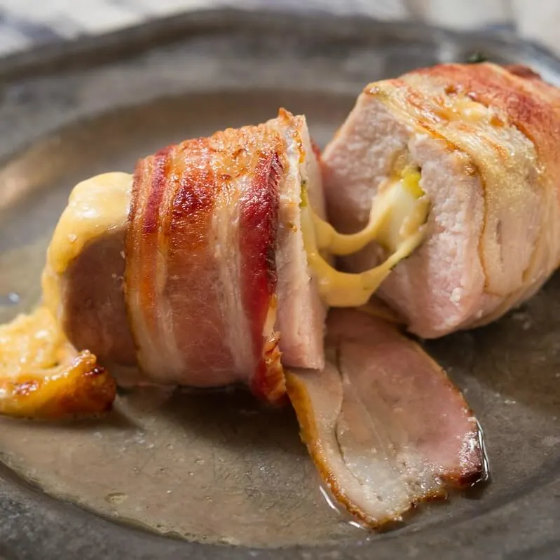 A metal plate holding a pork chop that\'s wrapped in bacon and stuffed with apple and cheese with the cheese pulled between the two halves. 
