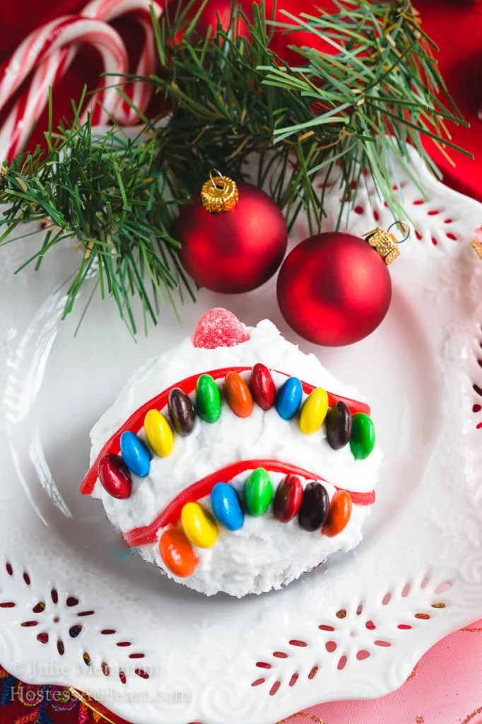 A cake ball frosted in white frosting decorated with candy pieces to resemble a Christmas bulb on a white snowflake plate. A sprig of greenery and two red Christmas bulbs are at the top of the plate.