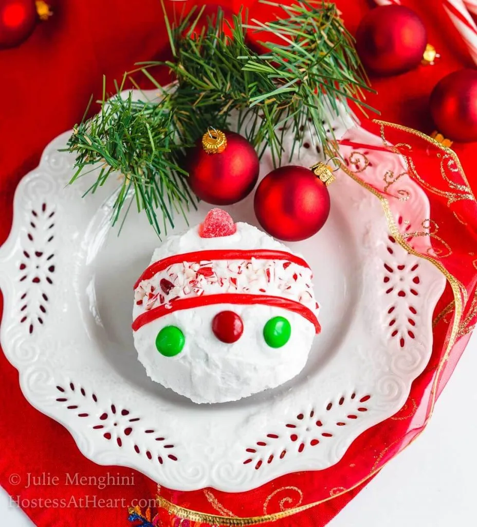 A cake ball frosted in white frosting decorated with candy pieces to resemble a Christmas bulb on a white snowflake plate. A sprig of greenery and two red Christmas bulbs are at the top of the plate.