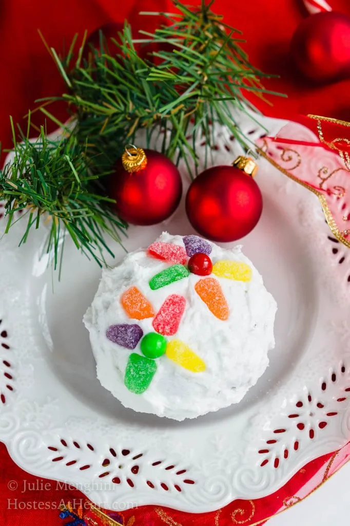 A cake ball frosted in white frosting decorated with candy pieces to resemble a Christmas bulb on a white snowflake plate. A sprig of greenery and two red Christmas bulbs are at the top of the plate.