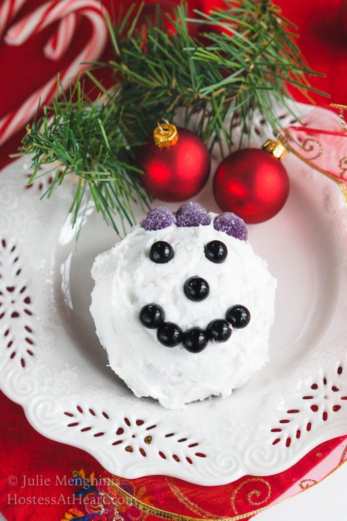 A cake ball frosted in white frosting decorated with candy pieces to resemble a snowman face on a white snowflake plate. A sprig of greenery and two red Christmas bulbs are at the top of the plate.
