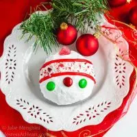 A cake ball frosted in white frosting decorated with candy pieces to resemble a Christmas bulb on a white snowflake plate. A sprig of greenery and two red Christmas bulbs are at the top of the plate.