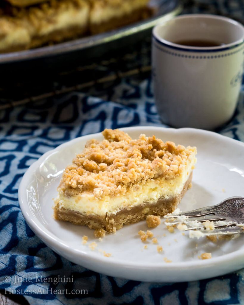 Side view of a bar of Cheesecake Cookie bars showing a graham cracker crust, cream cheese filling, and crumble top that\'s been cut with a fork also on a white plate. A cup of coffee sits in the background next to the pan of cookie bars.