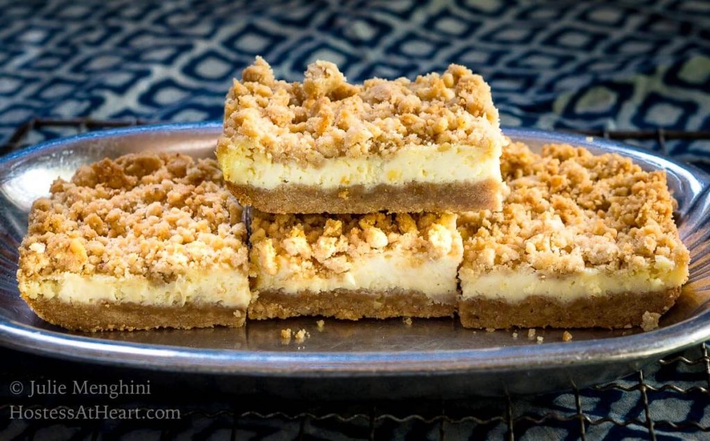 Side view of a stack of Cheesecake Cookie bars showing a graham cracker crust, cream cheese filling, and crumble top on a silver platter over a blue napkin. 