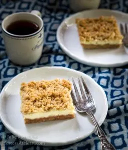 Top-down view of a bar of Cheesecake Cookie bars showing a graham cracker crust, cream cheese filling, and crumble top on a white plate. A cup of coffee sits in the background next to another plate will a cookie bar on it.