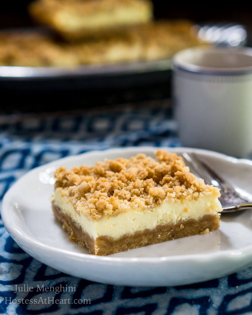 Side view of a bar of Cheesecake Cookie bars showing a graham cracker crust, cream cheese filling, and crumble top on a white plate. A cup of coffee sits in the background next to another plate will a cookie bar on it.