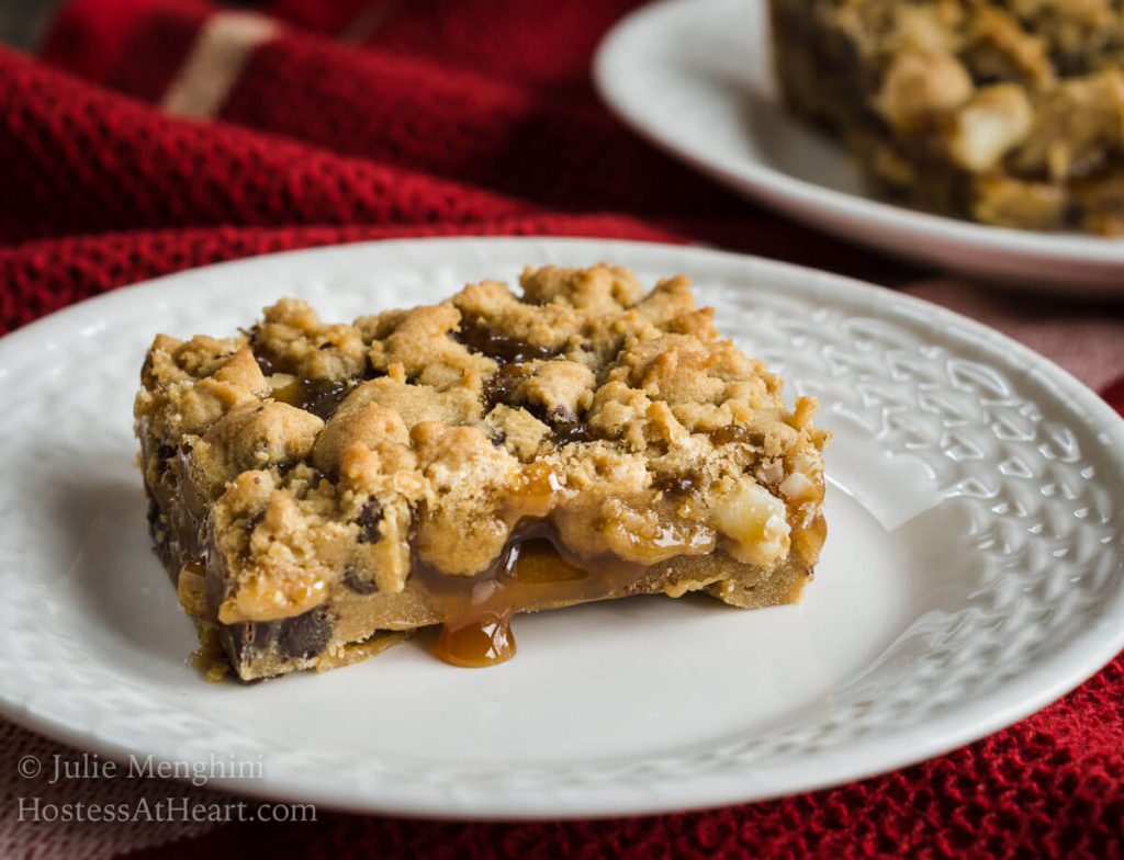 Slice of Chocolate Caramel Cookie Bar over a white plate with a drop of caramel running out of it. A second plate sits behind it over a red napkin.