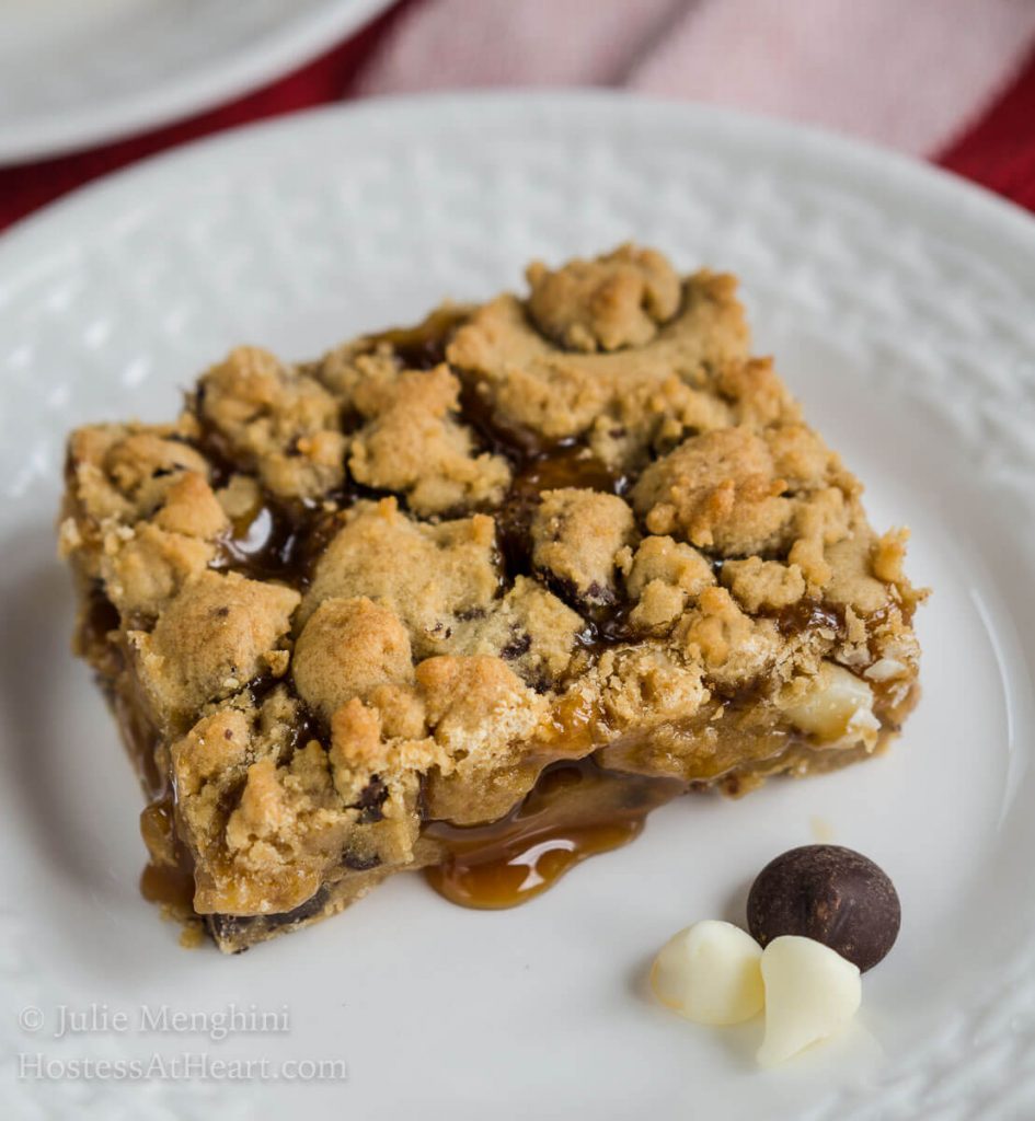 Slice of Chocolate Caramel Cookie Bar over a white plate with a drop of caramel running out of it and the chocolate chips sitting next the bar.