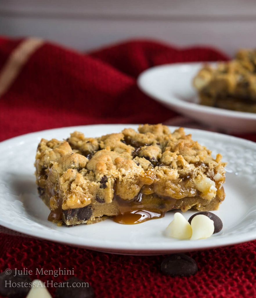 Slice of Chocolate Caramel Cookie Bar over a white plate with a drop of caramel running out of it and the chocolate chips sitting next to the bar over a red napkin. A second plate sits in the background.