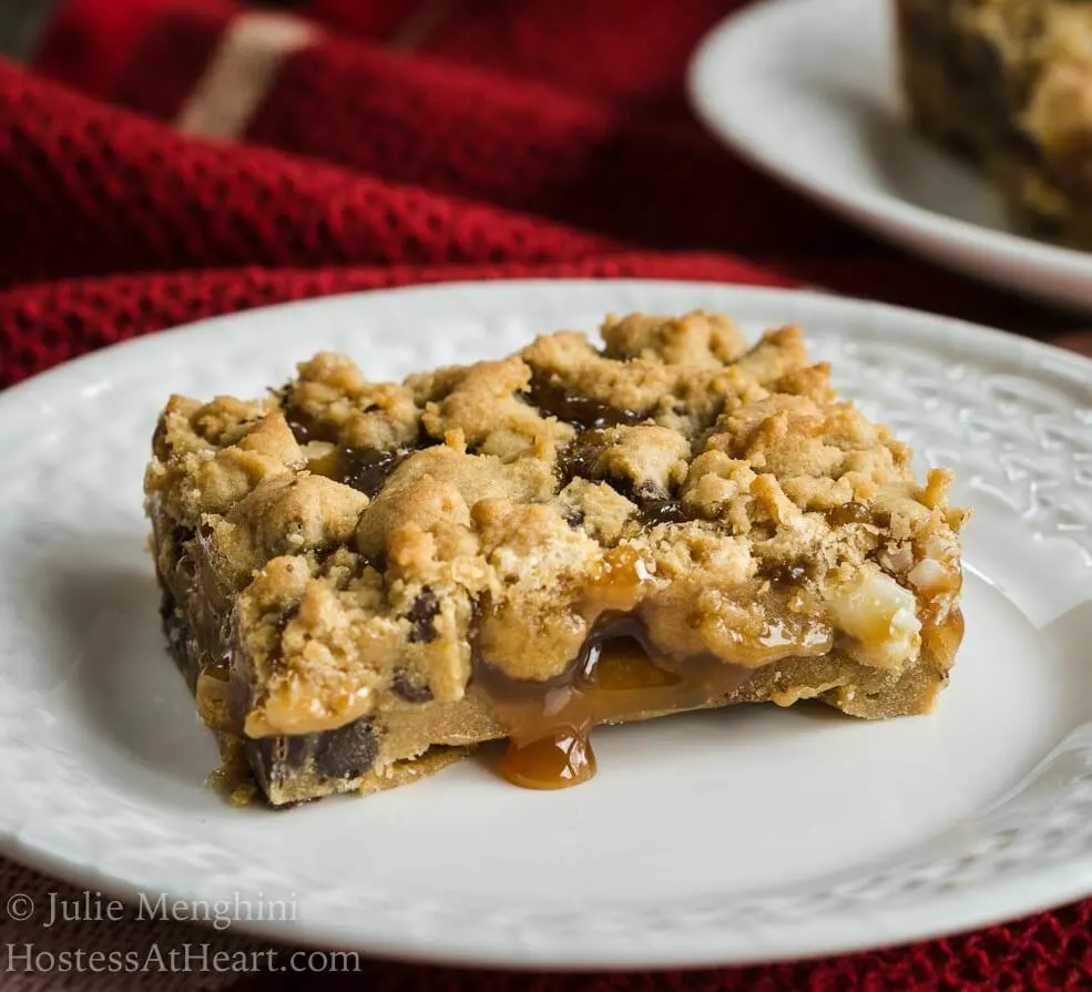 A piece of Chocolate caramel cookie bar on a white plate. It has a crumbled top and a drip of caramel running onto the plate. A second plate with a bar on it sits in the background over a red napkin.