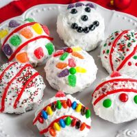 Cake balls frosted in white frosting decorated with candy pieces to resemble Christmas bulbs on a white snowflake plate sitting over a red napkin.
