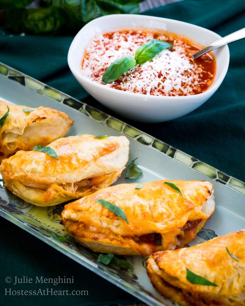 A tray of Cheese and Mushroom Empanadas sitting in front of a white dish of tomato marinara garnished with grated cheese and fresh basil.