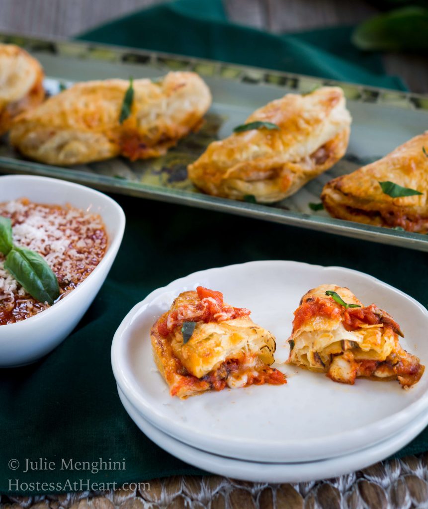 A tray of Cheese and Mushroom Empanadas sitting behind a plate of topped with a sliced empanada on a white plate next to a bowl of tomato marinara garnished with grated cheese and fresh basil.