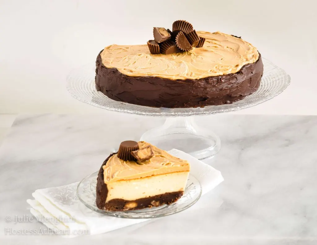 A brownie cheesecake with a smooth chocolate edge and shaved chocolate over the top sits on a clear glass cake stand. A piece of the chocolate cheesecake sits below it over a white napkin.