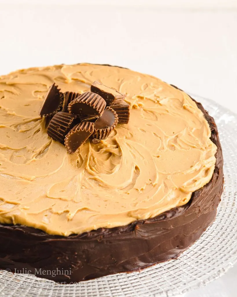 Top angled view of a Close up of a brownie cheesecake with a chocolate edge, a peanut butter topping, and peanut butter cups as a garnish sits on a clear glass cake stand.