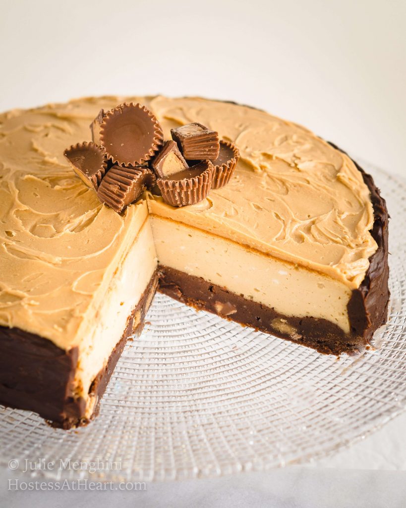 Top angled view of a Close up of a brownie cheesecake with a brownie crust, cream cheese filling, and a peanut butter topping and peanut butter cup garnish sits on a clear plate on a glass cake stand.