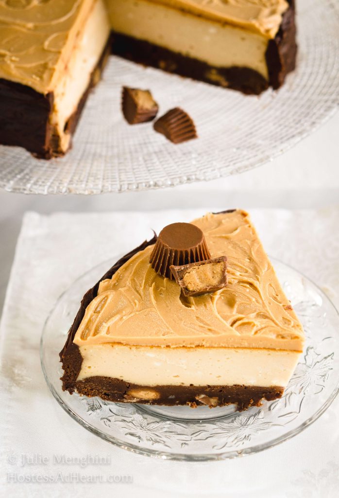 Close up of a slice of cheesecake with a brownie crust, cream cheese filling and a peanut butter topping and peanut butter cup garnish sits on a clear plate over a white napkin.