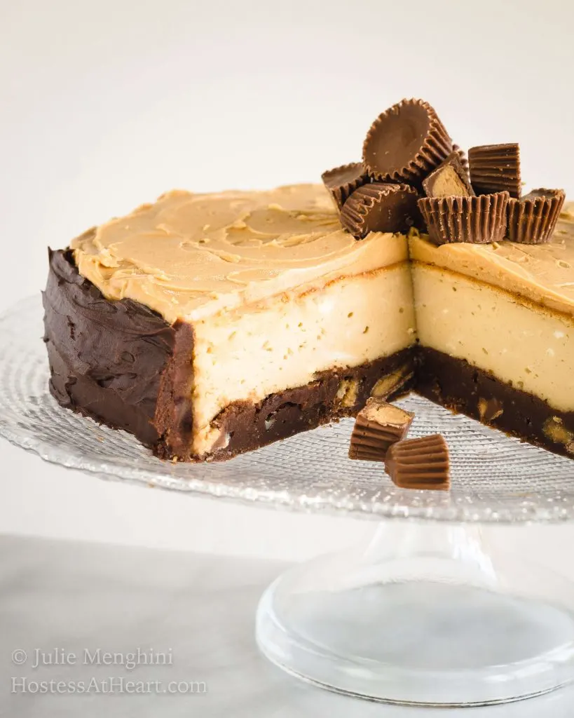 Side view of a Close up of a brownie cheesecake with a brownie crust, cream cheese filling and a peanut butter topping and peanut butter cup garnish sits on a clear plate on a glass cake stand.