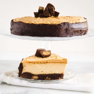 Side view of a cake stand holding a Cheesecake with a smooth chocolate edge and large shaved pieces of chocolate on the top. A slice of the cheesecake sits below it on a clear glass plate over a white napkin.