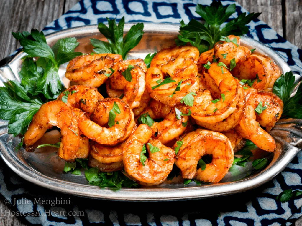 45 degree angle photo of red colored blackened shrimp on a silver platter garnished with parsley and sitting on a blue checked napkin.