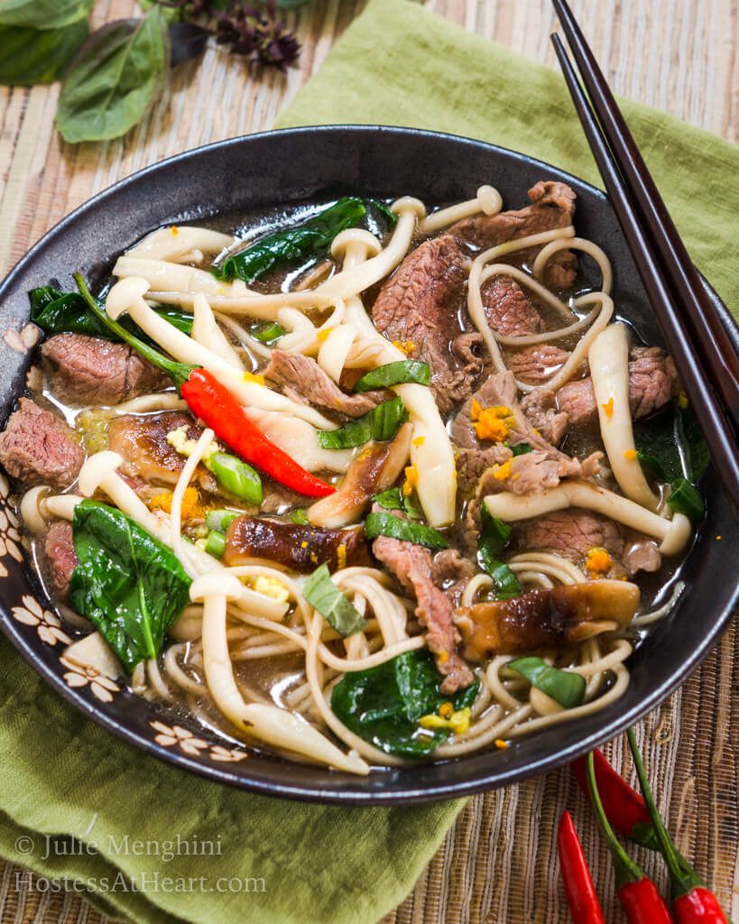 Top-down view of a dark gray bowl filled with thin slices of beef, mushrooms, buckwheat noodle, Thai basil and garnished with spicy red peppers over a green napkin. A set of chopsticks sits on the back of the bowl and red peppers sit in front of the bowl.