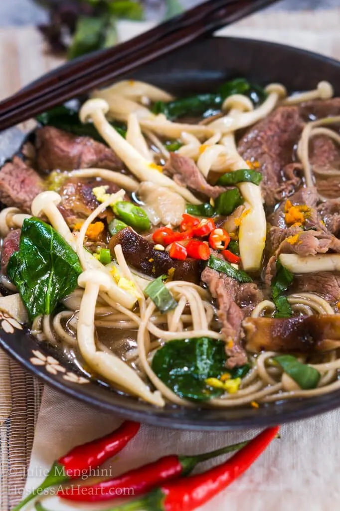 Top-down view of a dark gray bowl filled with thin slices of beef, mushrooms, buckwheat noodles, Thai basil and garnished with spicy red peppers. A set of chopsticks sits on the back of the bowl and red peppers sit in front of the bowl.