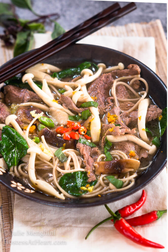Top-down view of a dark gray bowl filled with thin slices of beef, mushrooms, buckwheat noodles, Thai basil and garnished with spicy red peppers. A set of chopsticks sits on the back of the bowl and red peppers sit in front of the bowl. Additional Thai basil sits in the background.