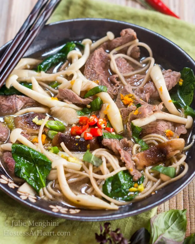 Top-down view of a dark gray bowl filled with thin slices of beef, mushrooms, buckwheat noodles, Thai basil and garnished with spicy red peppers. A set of chopsticks sits on the back of the bowl and Thai basil sits in front of the bowl.
