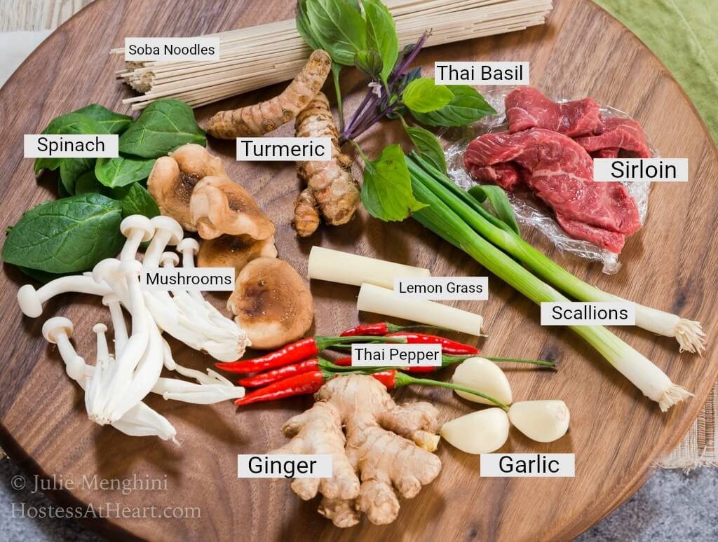 A wooden cutting board filled with the ingredients used in an Asian Beef Noodle Bowl including Thai Peppers, ginger, garlic, mushrooms, beef, green onions, turmeric, soba noodles, and spinach.