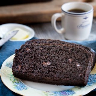 Two slices of chocolate banana bread showing melted chocolate chips in the crumb on a floral plate over a blue napkin. A cup of espresso sits behind it with a pad of butter in a small white dish with a knife next to it.