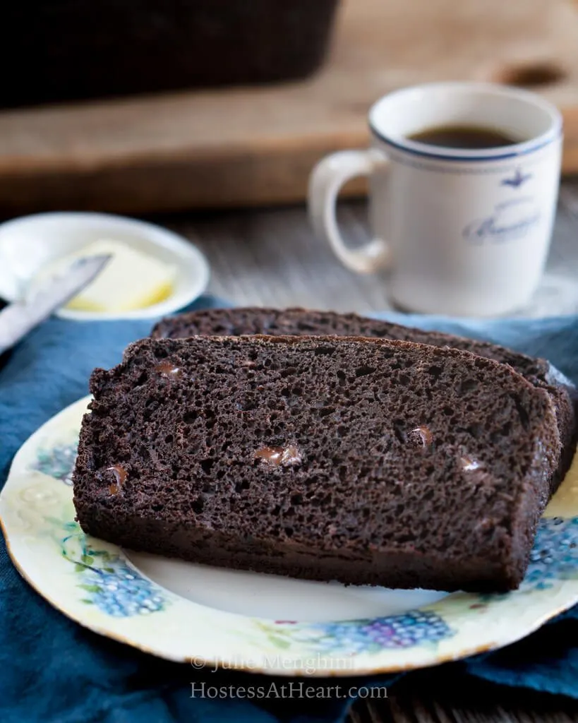 Two slices of chocolate banana bread showing melted chocolate chips in the crumb on a floral plate over a blue napkin. A cup of espresso sits behind it with a pad of butter in a small white dish with a knife next to it.