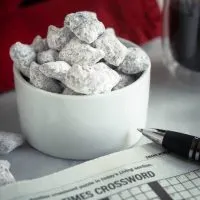A close up of a white ramekin filled with powdered sugar covered peanut butter pretzels. A crossword puzzle with a pen and a cup of coffee sit next to the bowl. A red napkin sits in the background.
