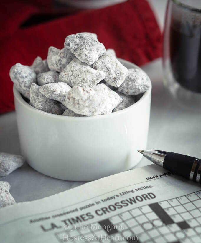 A close up of a white ramekin filled with powdered sugar covered peanut butter pretzels. A crossword puzzle with a pen and a cup of coffee sit next to the bowl. A red napkin sits in the background.