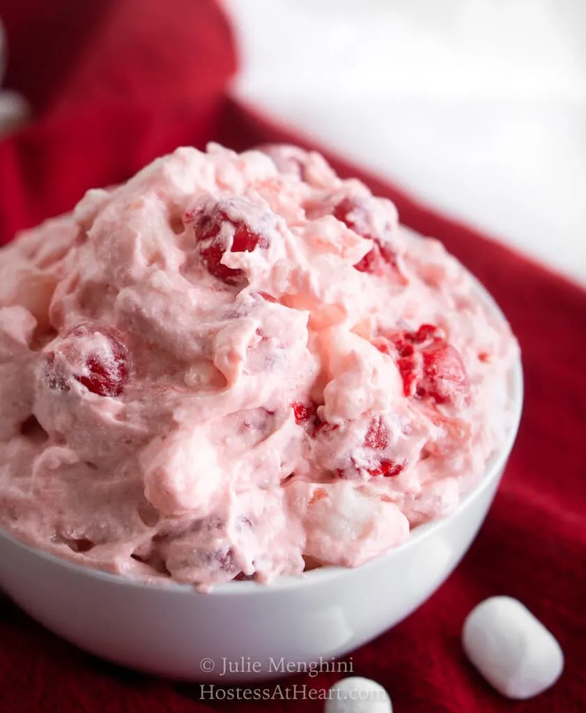 Close up top view of a white bowl filled with a creamy pink Cherry fluff salad sitting on a red napkin with mini marshmallows scattered around the bowl.