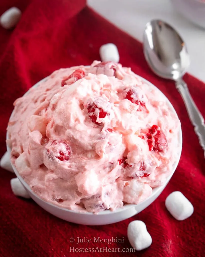 Top-down photo of a white bowl of pink cherry fluff on a red napkin with mini marshmallows scattered in front. 