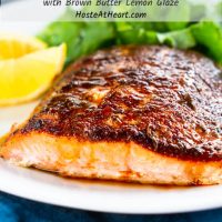 A close up of a white plate holding a piece of baked Salmon with blackened seasoning and a drizzle of a brown butter lemon drizzle. Sliced lemon and a lettuce salad sit in the background.  The title 