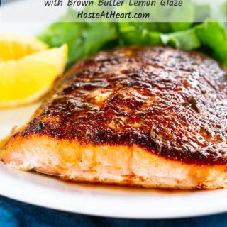 A close up of a white plate holding a piece of baked Salmon with blackened seasoning and a drizzle of a brown butter lemon drizzle. Sliced lemon and a lettuce salad sit in the background.  The title "Baked Blackened Salmon with Brown Butter Lemon Glaze" runs across the top.